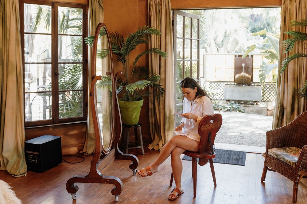 Woman sitting on chair in bohemian room with floor mirror, tropical plants, and French doors leading to garden.