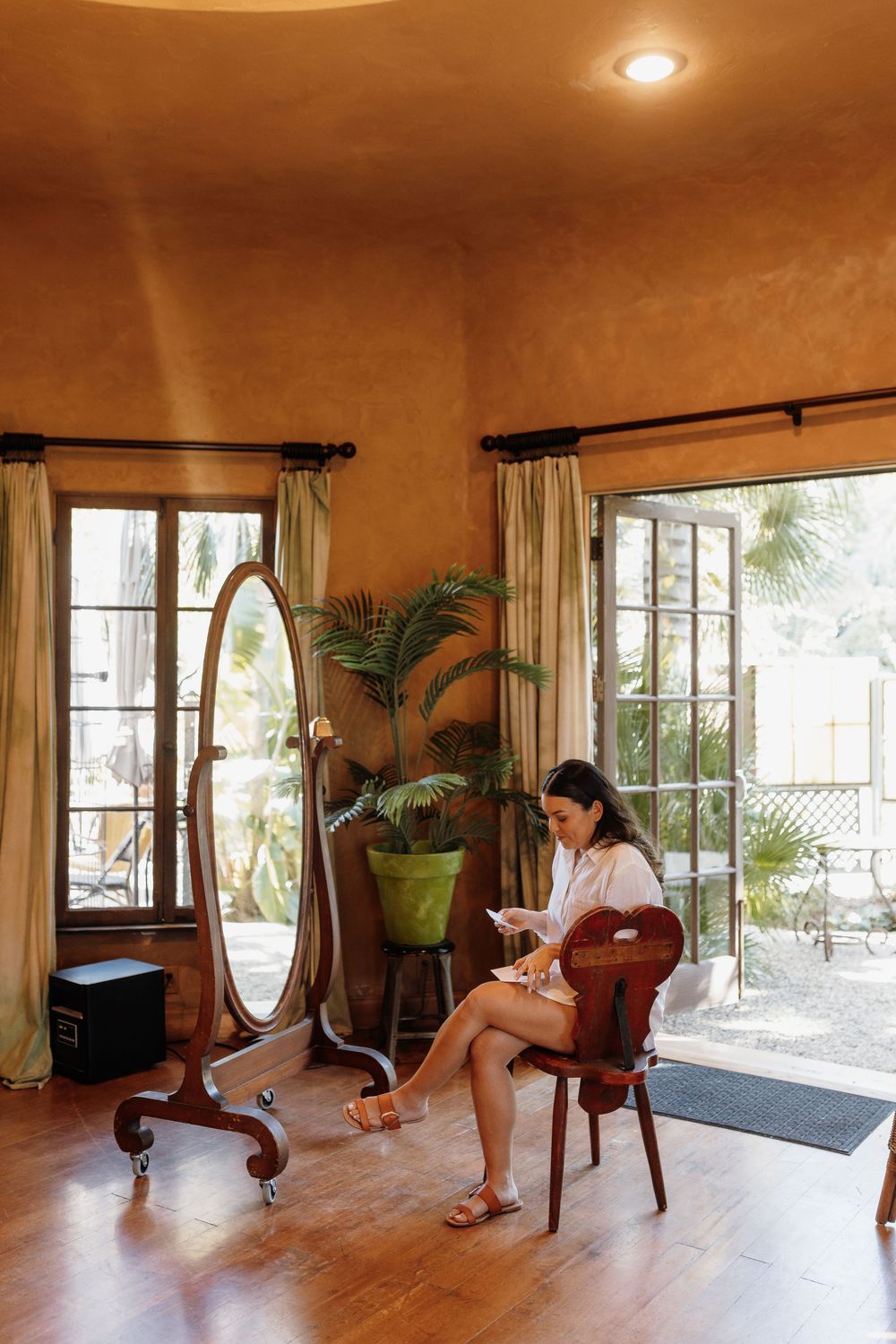 Woman reading in a warm-toned room with a cheval mirror, tropical plants, and French doors leading to a sunny patio.