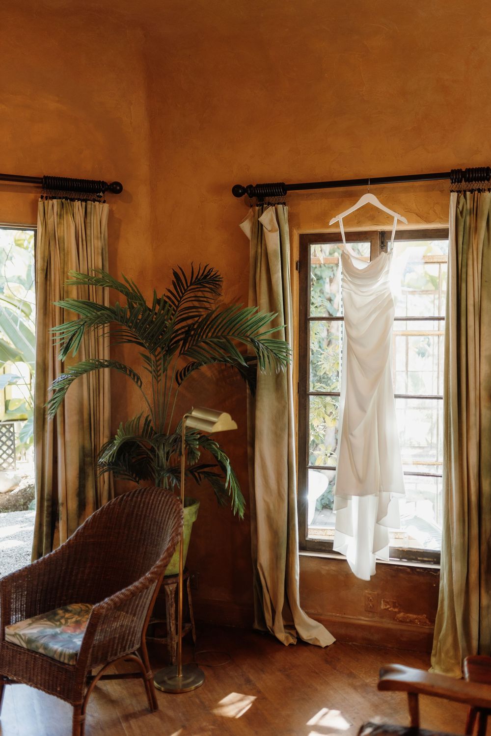White wedding dress hanging on a curtain rod in a warm-toned room with wicker chair and tropical plant.
