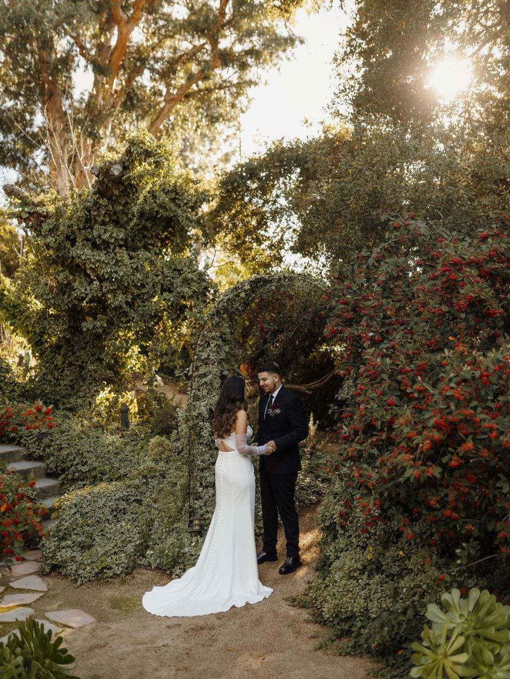 Bride and groom share a romantic moment under a lush garden arch with red flowers and golden sunlight.