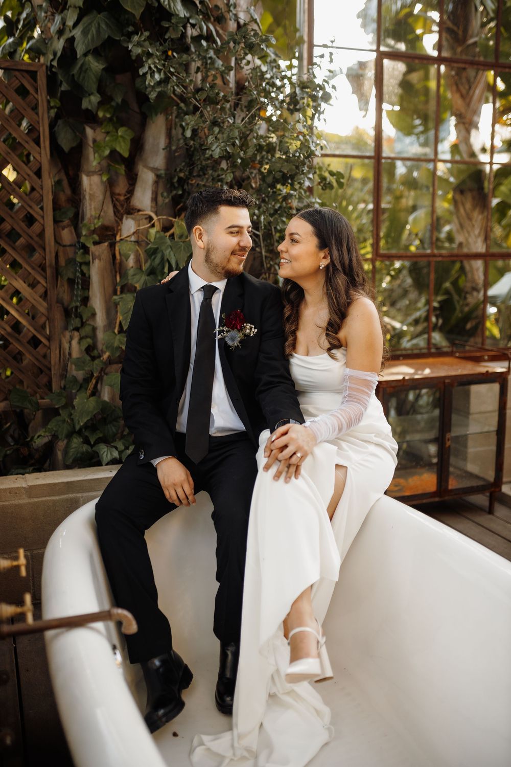 A bride and groom sit together on a vintage bathtub, smiling at each other, surrounded by lush greenery indoors.
