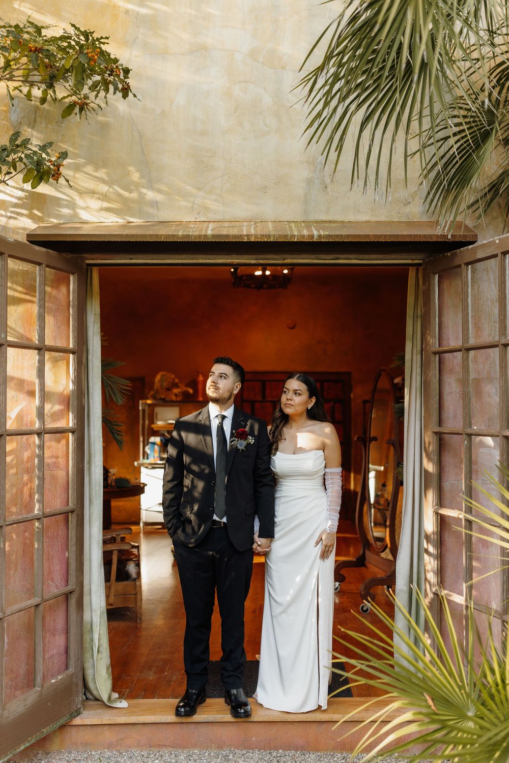 Bride in white gown and groom in black tuxedo stand together in an elegant doorway at their wedding venue.