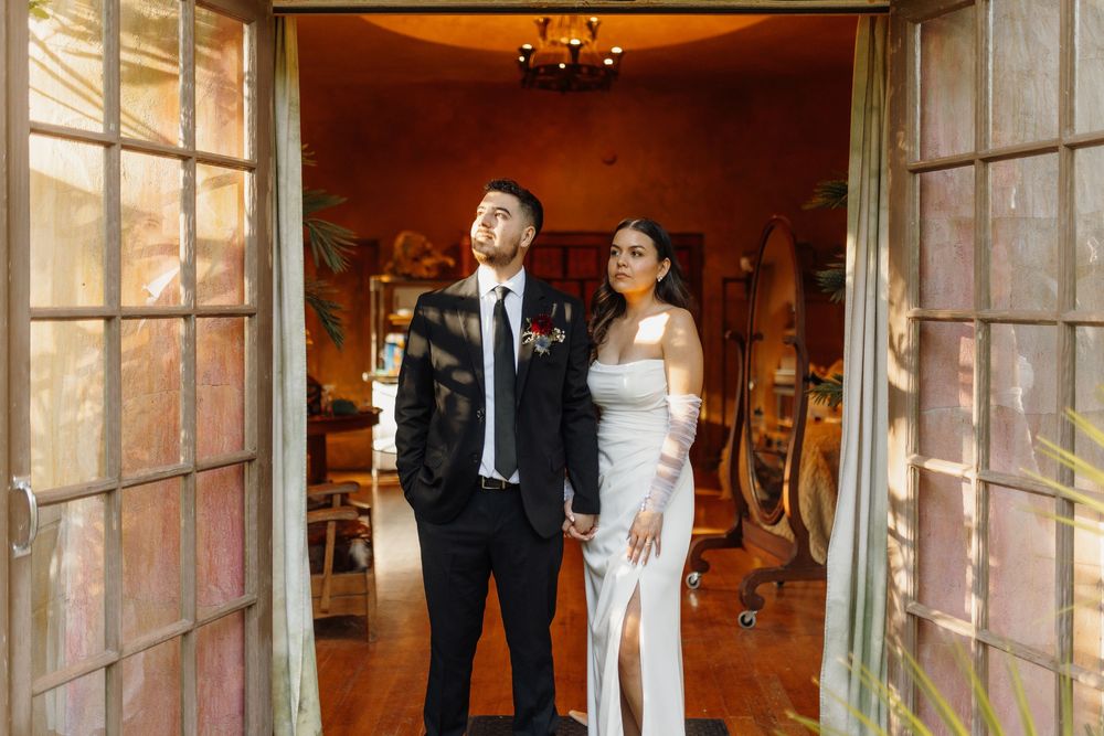 Elegant bride and groom standing in ornate doorway, wearing white gown and black suit with floral boutonniere.