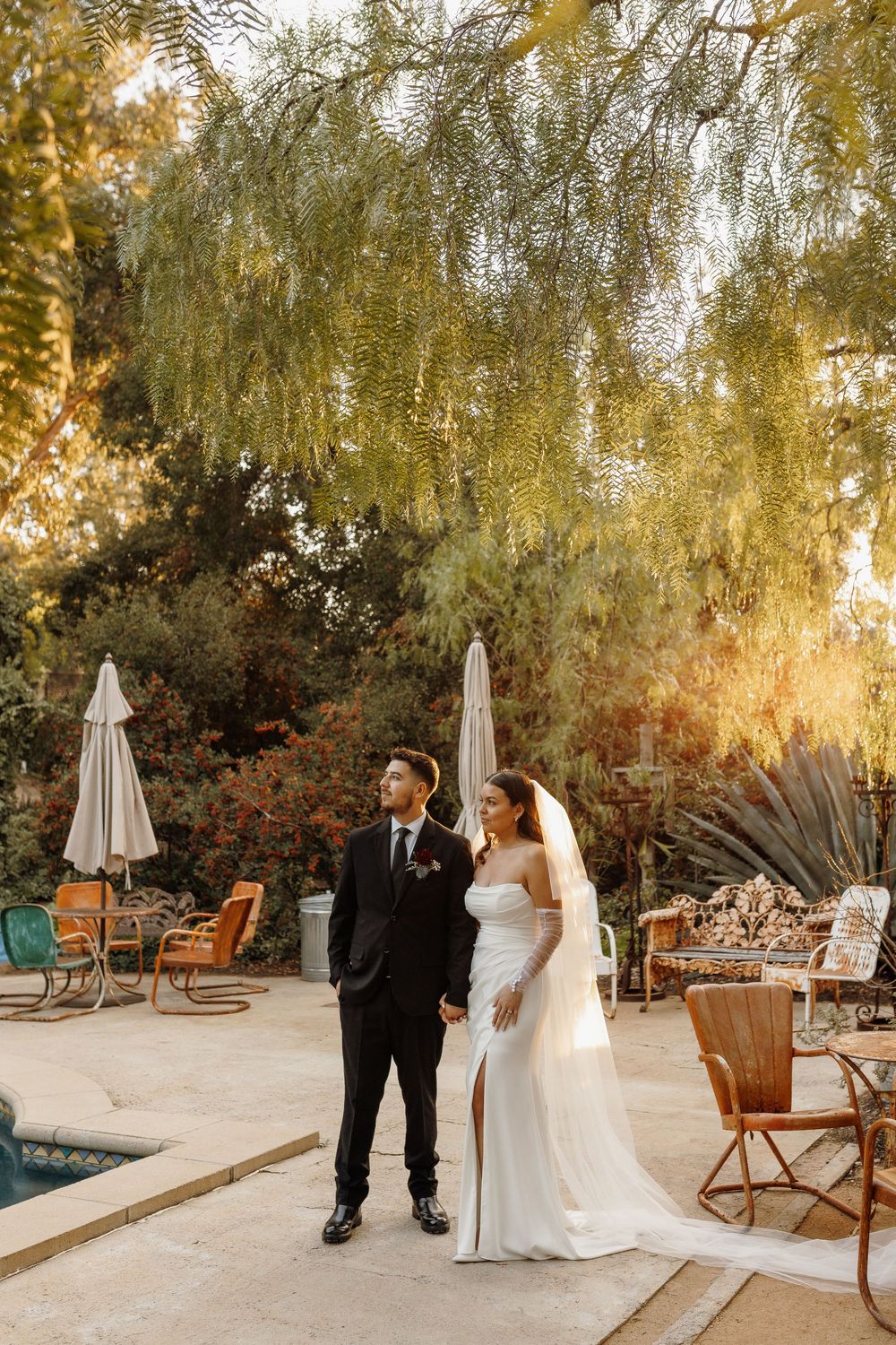 Bride in white gown and veil stands with groom in black suit by poolside garden at golden hour wedding.