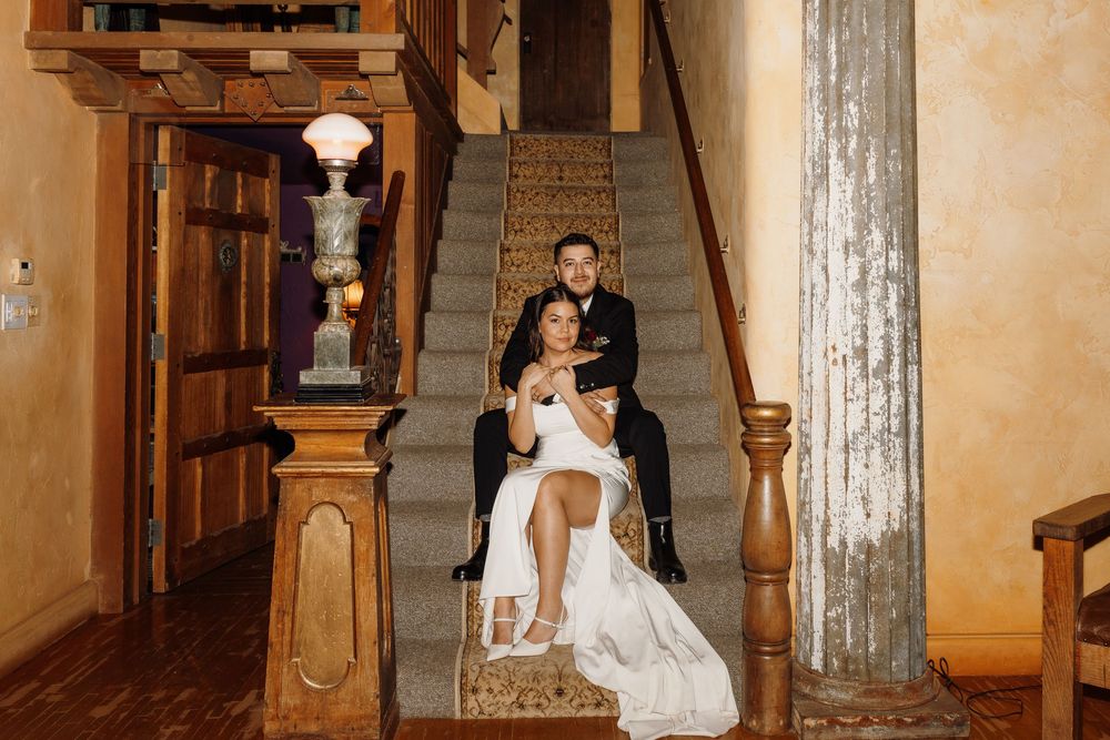 Bride and groom pose elegantly on wooden staircase in rustic venue, bride seated in groom's lap in wedding attire.
