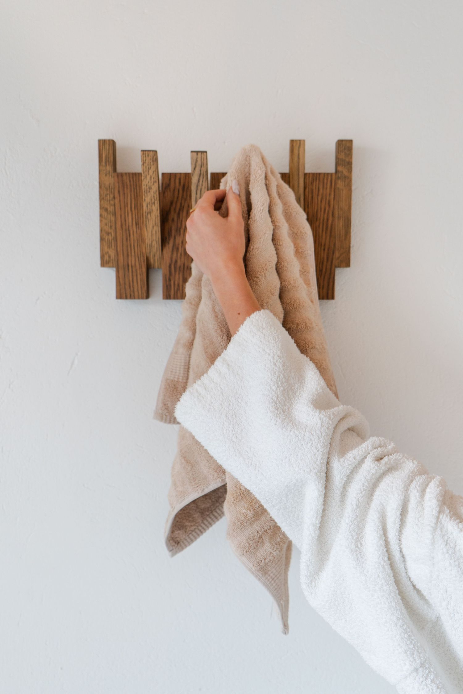 Curated product photography of a hand in white robe gown hanging a beige towel onto a wooden wall-mounted hook rack.