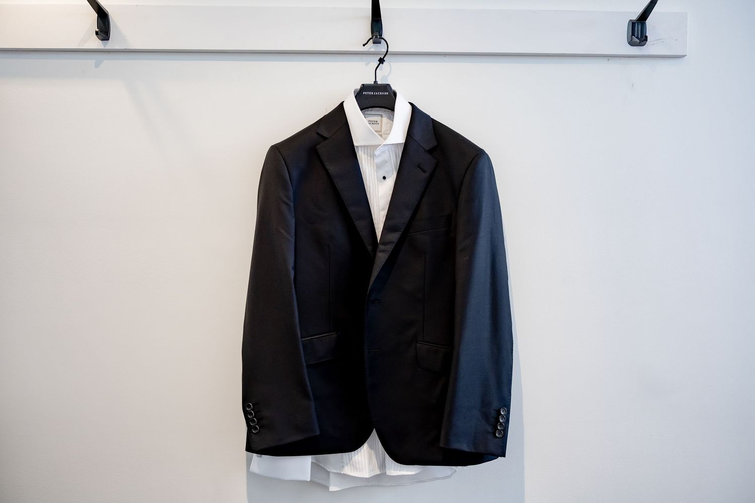Black suit jacket with white dress shirt on a hanger displayed against a white wall.