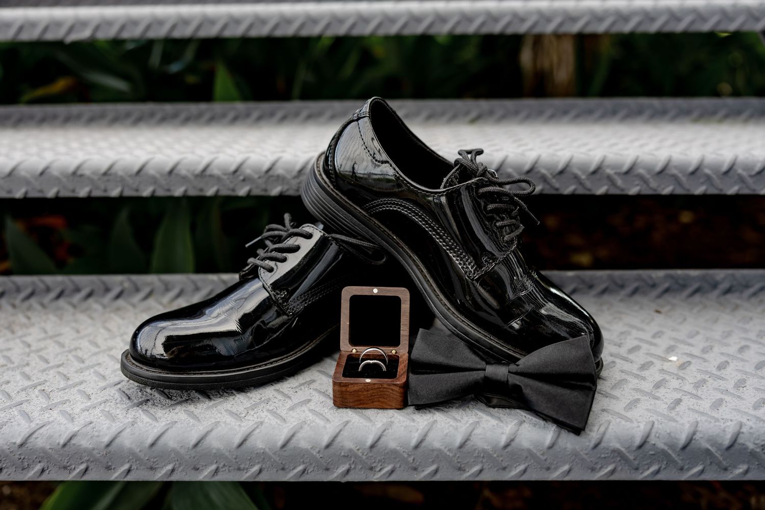 Shiny black patent leather dress shoes with a bow tie and wooden ring box on a metal diamond plate surface.