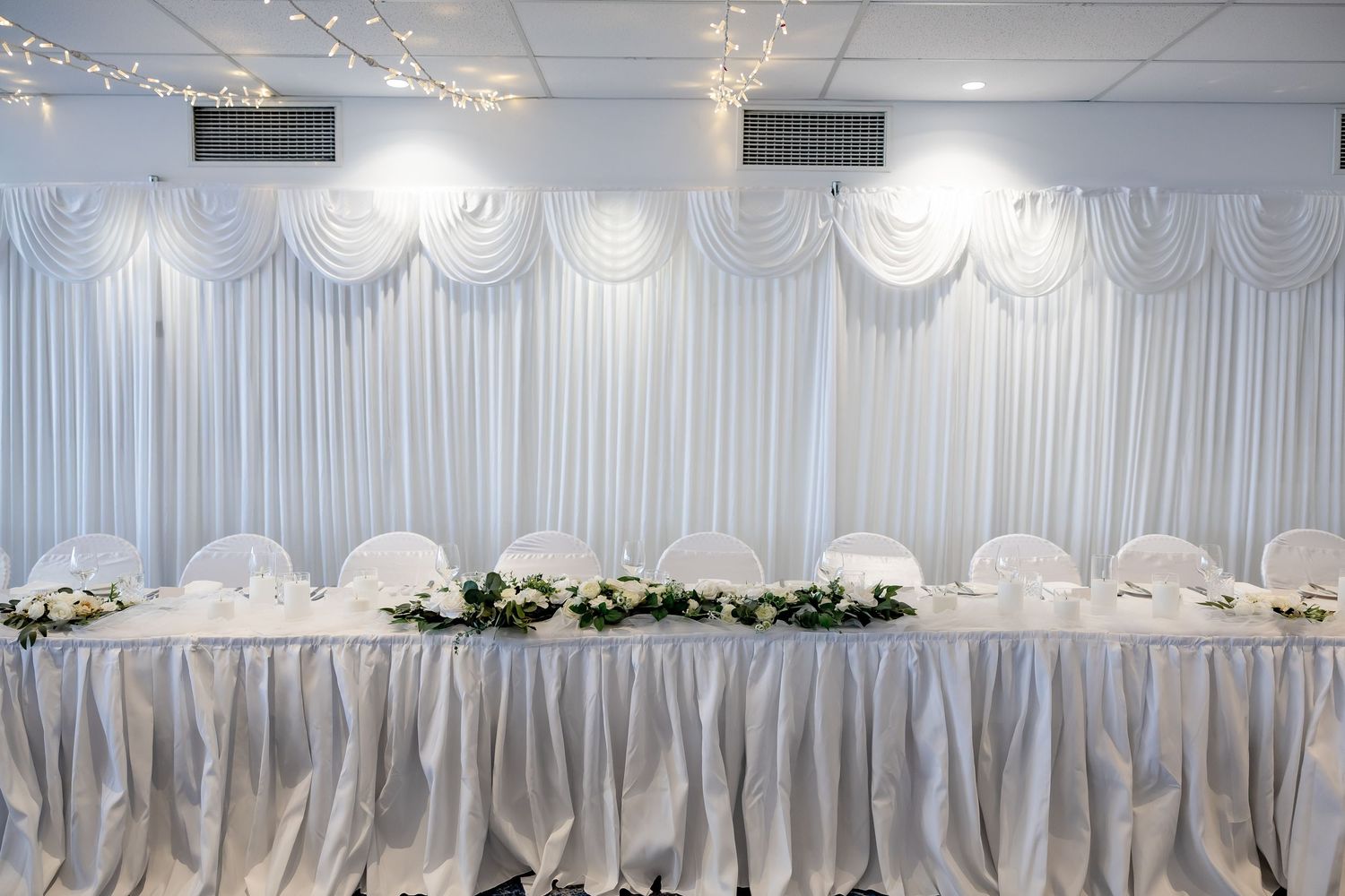 Elegant white wedding head table with floral centerpiece, draped fabric backdrop, and fairy lights in a banquet hall.