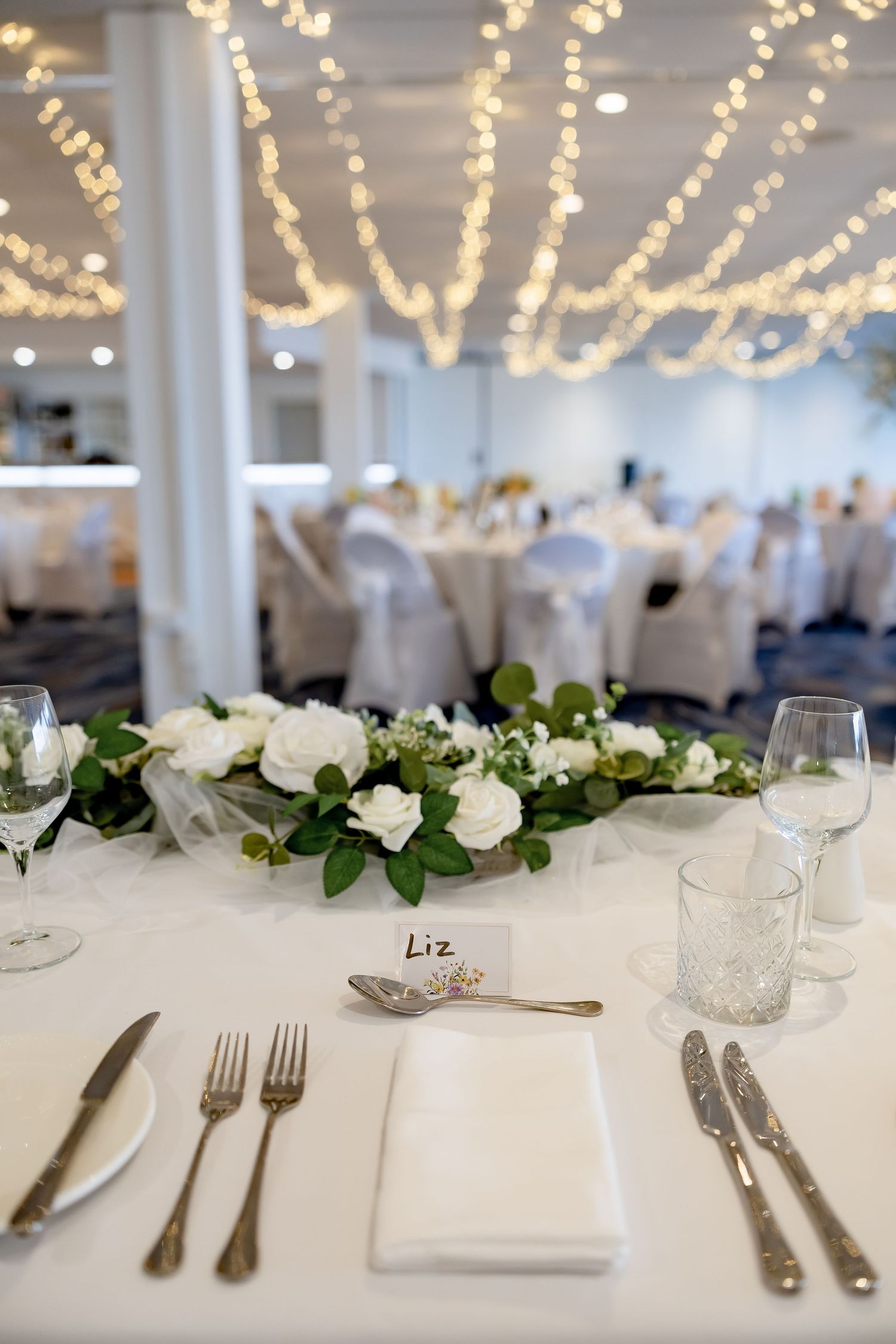 Elegant wedding reception table with white rose centerpiece, place card reading Liz, silver cutlery, and fairy lights above.