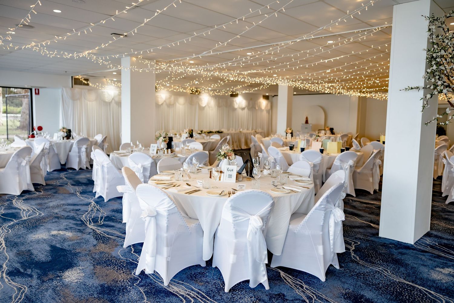 Romantic wedding reception hall with fairy lights, white chair covers, and round tables on blue carpet.