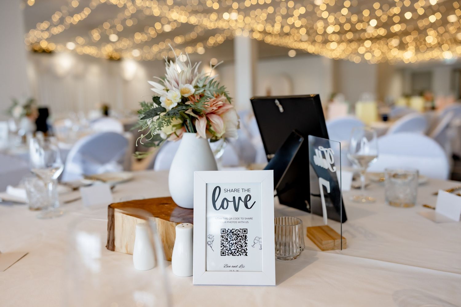 Elegant wedding reception table with floral centerpiece, QR code love sign, and twinkling fairy lights in background.