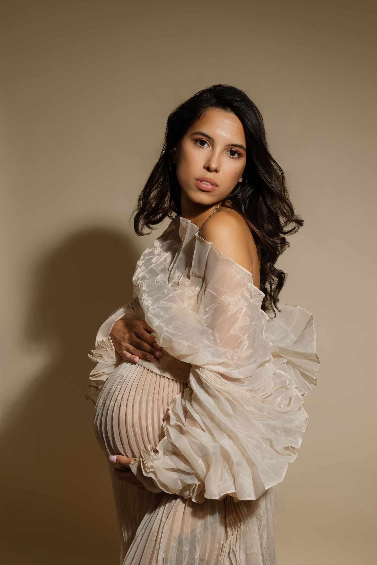 Pregnant woman in elegant ruffled sheer cream gown cradling baby bump in a soft beige studio portrait.