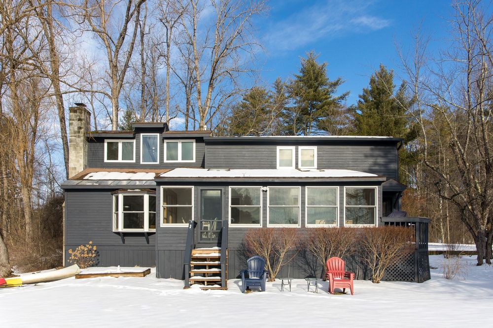 Dark gray cottage with sunroom covered in snow surrounded by bare winter trees with red chairs outside.