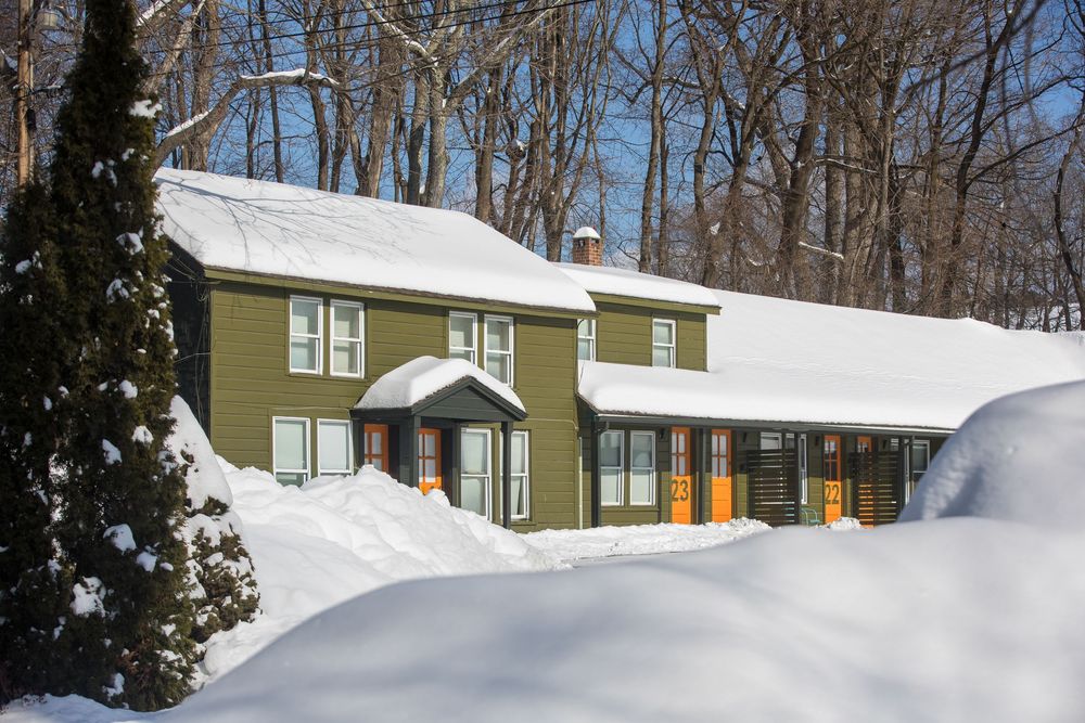 Green two-story colonial home buried in deep snow surrounded by tall bare trees in winter landscape.