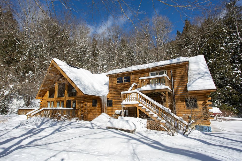 Rustic log cabin with large A-frame windows and exterior staircase covered in deep snow in winter.