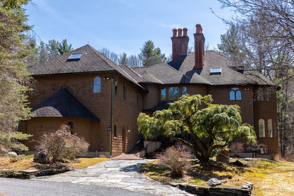 Large multi-story shingle-style home with chimneys and mature weeping tree in wooded setting.