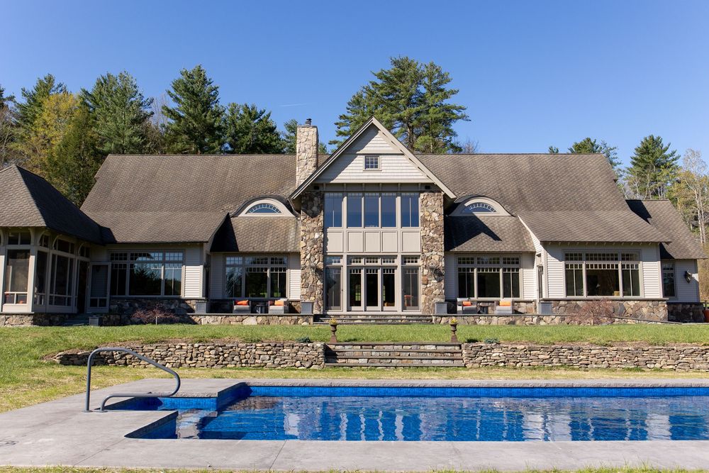 Luxury stone and shingle home with pool in foreground surrounded by tall trees under clear blue sky.