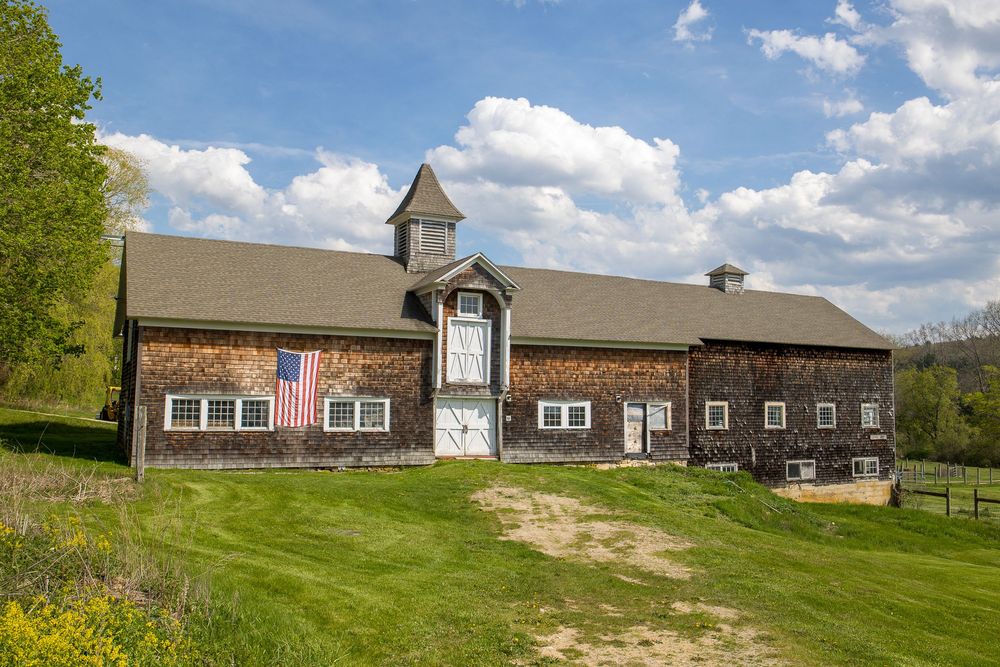 Rustic barn with American flag, cupola, and green lawn set against a wooded hillside.