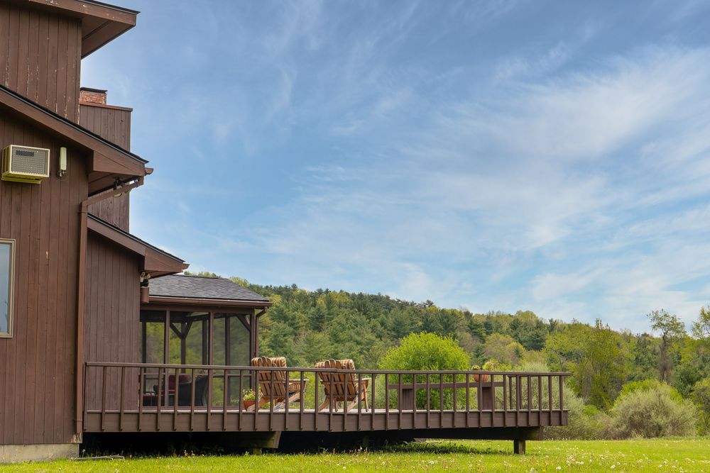 Wooden deck with outdoor chairs overlooking a green valley and forested hillside under blue sky.
