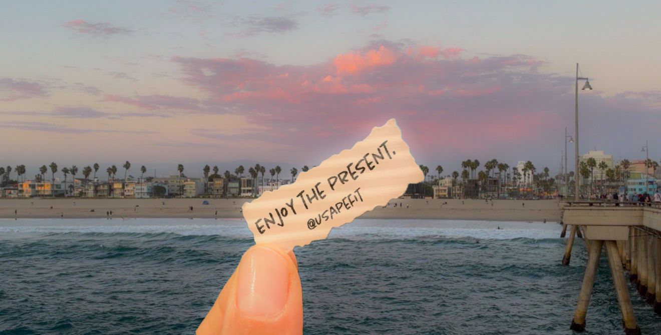 Hand holding a note saying Enjoy the Present at a scenic beach pier during sunset with palm trees.