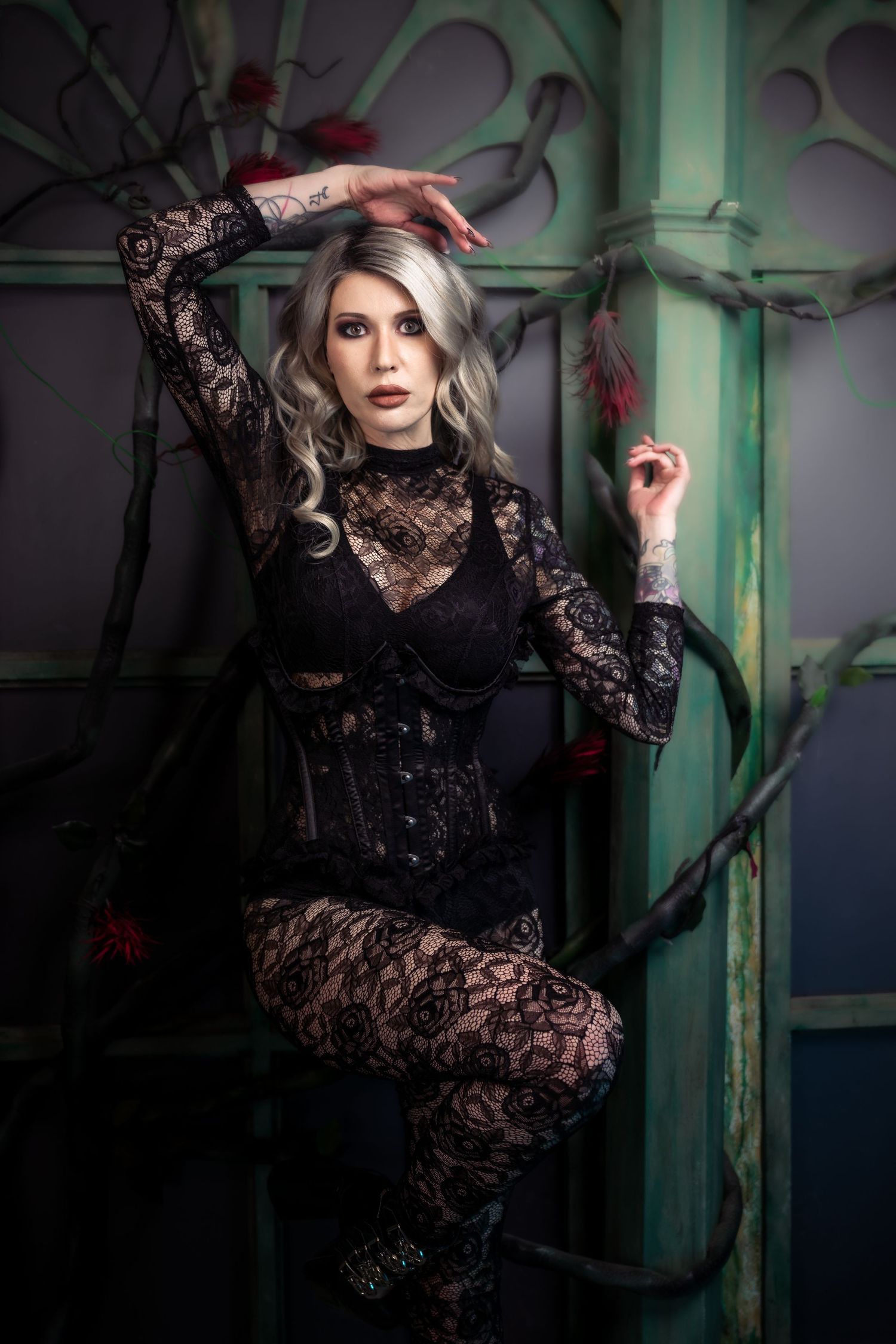 Woman in black lace gothic outfit with corset poses dramatically against a dark, moody backdrop with red accents.