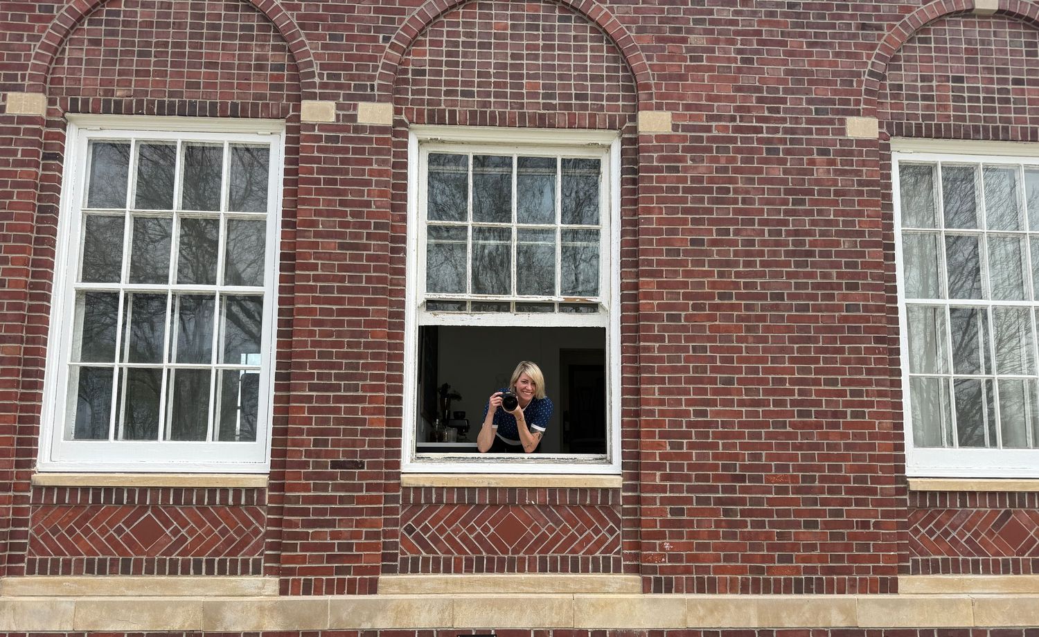 Person photographing from open window of red brick building with white-framed windows.