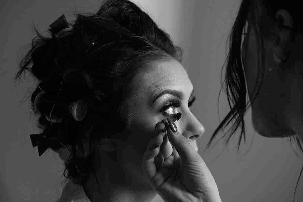 Black and white photo of a woman having false eyelashes applied by a makeup artist during bridal preparations prior to her Greenhill wedding
