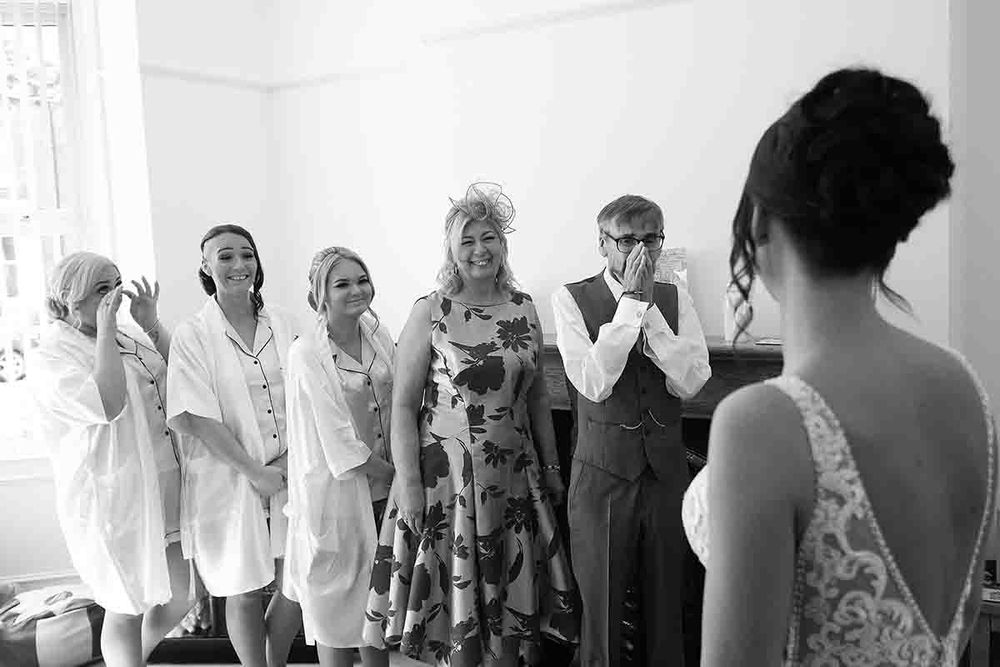 Black and white photo of a bride revealing her wedding dress to emotional family and friends in a bright room.