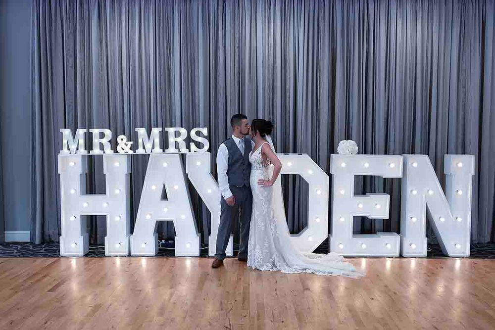 Bride and groom kissing in front of illuminated MR & MRS HAYDEN marquee letters on a wooden dance floor at Greenhill Hotel - Wigton