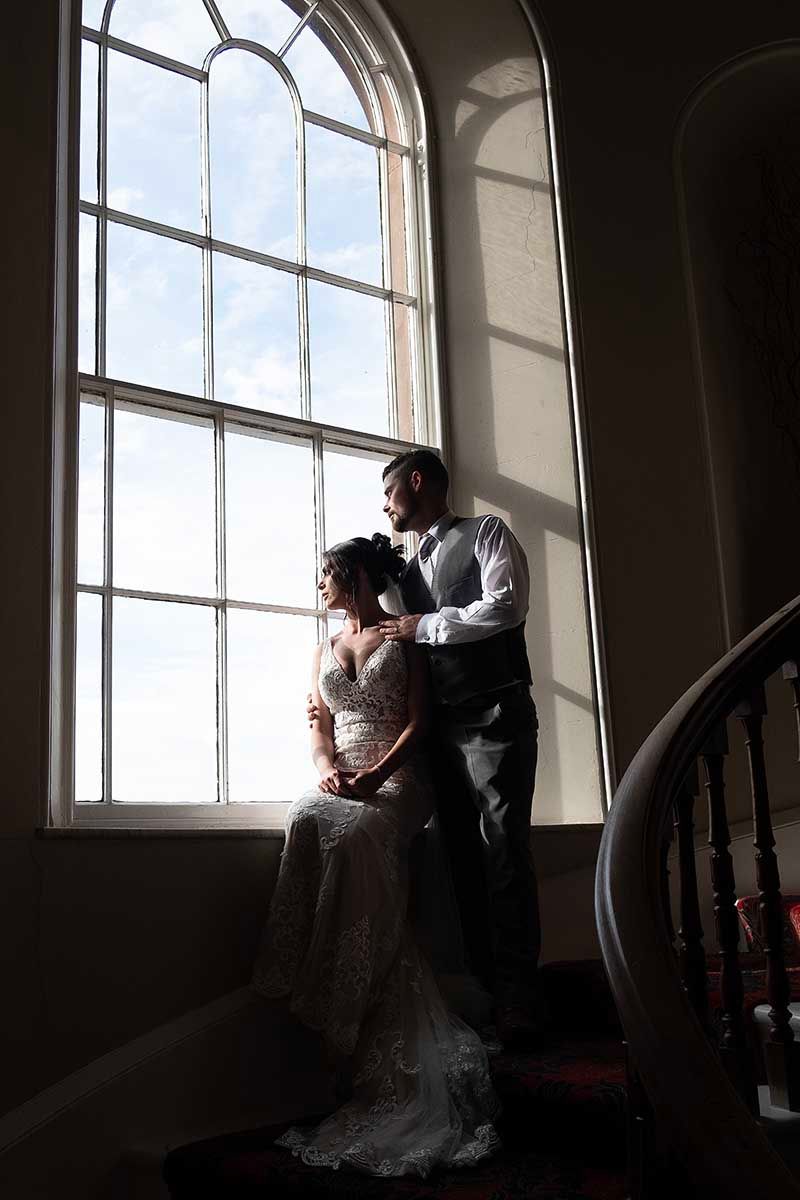 Bride and groom silhouetted by large arched window light in elegant venue, romantic wedding portrait at Greenhills iconic feature window