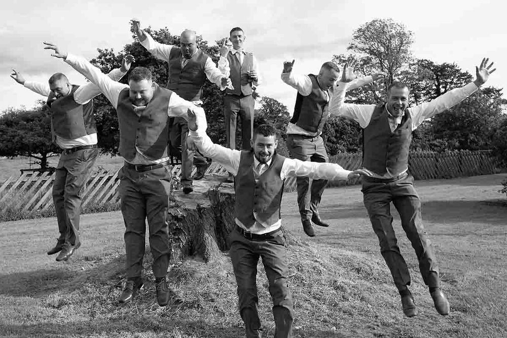 Black and white photo of groomsmen joyfully jumping in the air outdoors at a wedding celebration at Greenhill Hotel near Wigton