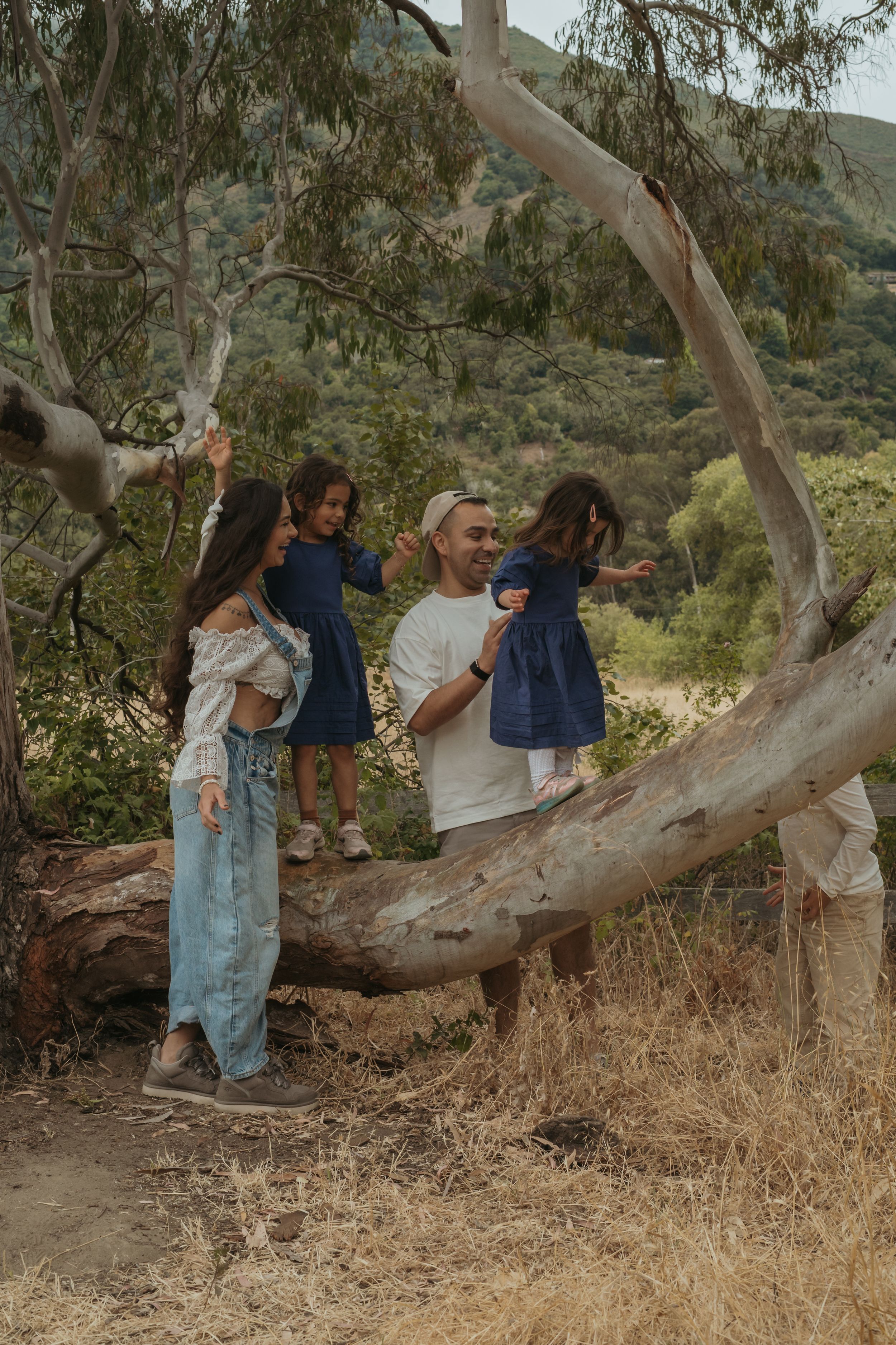 Family of four enjoying a nature walk, with two young girls in blue dresses climbing a fallen eucalyptus tree outdoors.