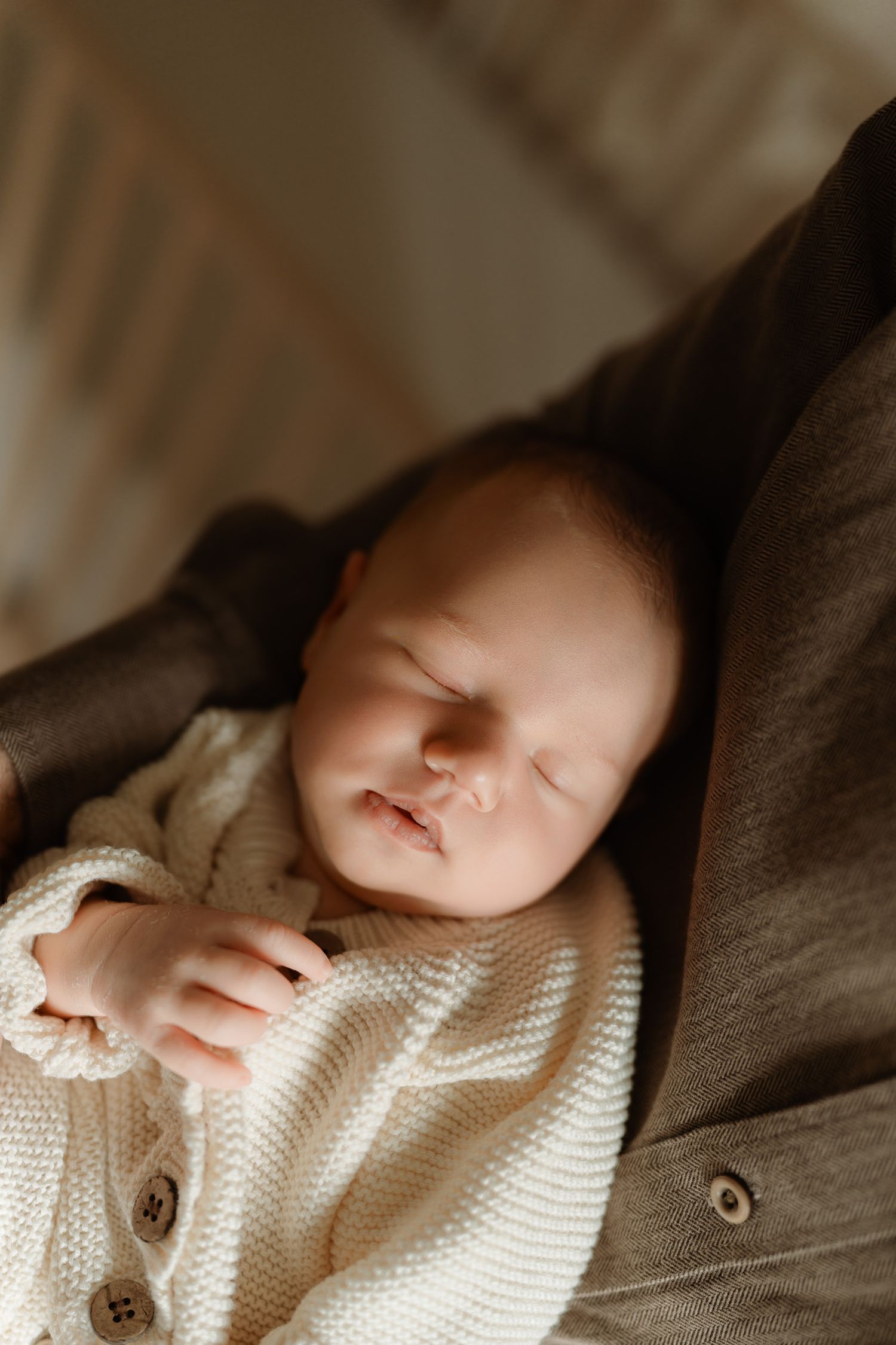 Sleeping newborn baby in cream knit sweater resting peacefully in adult's arms, soft warm lighting.