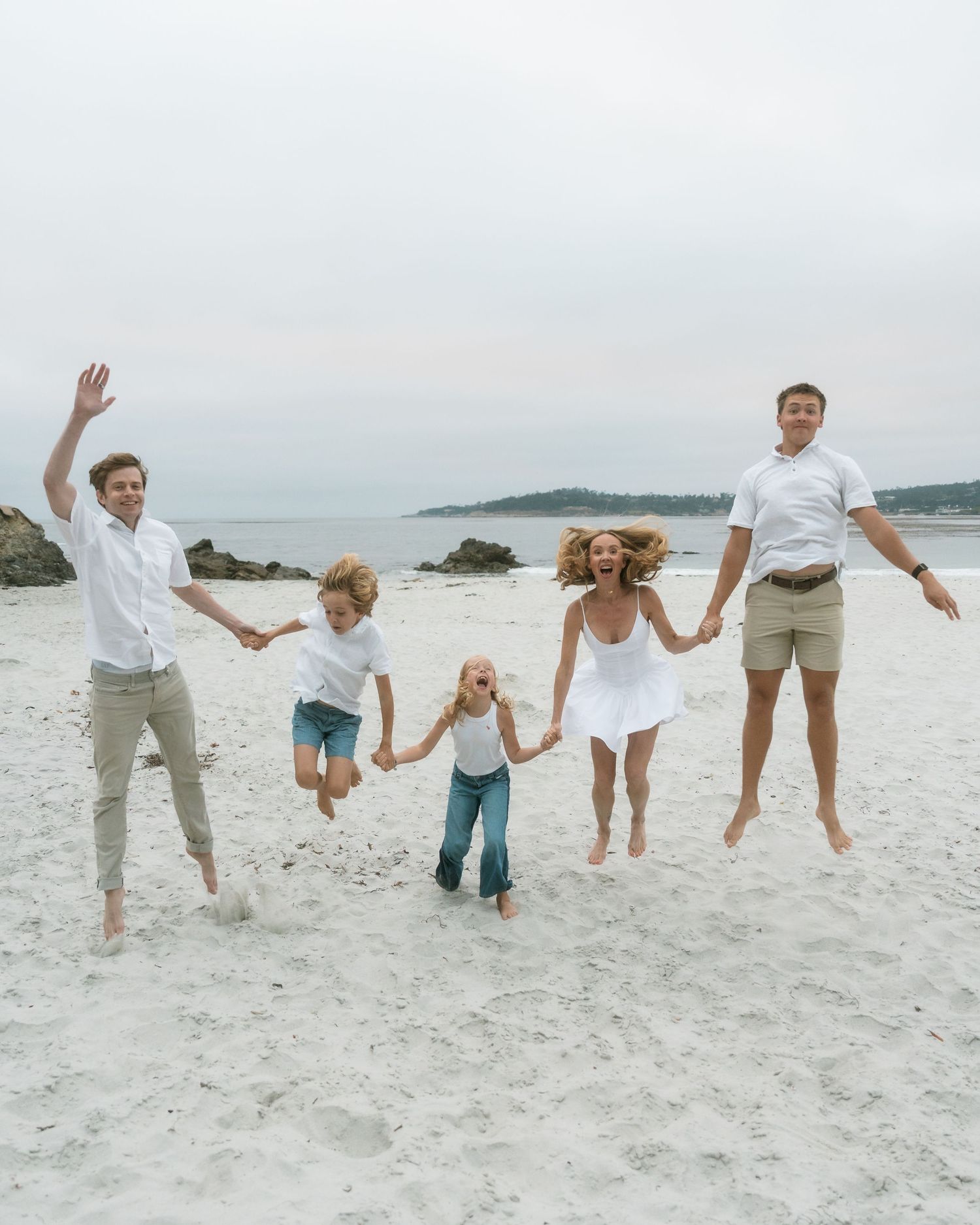 A joyful family of five holds hands and jumps together on a sandy beach wearing coordinated white outfits.
