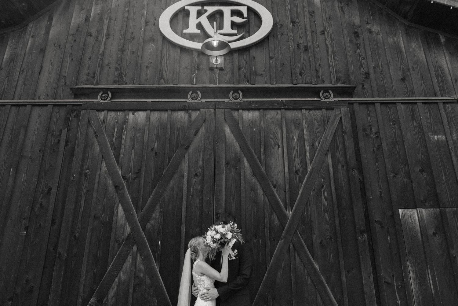 Black and white photo of a bride holding a bouquet in front of a rustic wooden barn door with KF monogram sign.