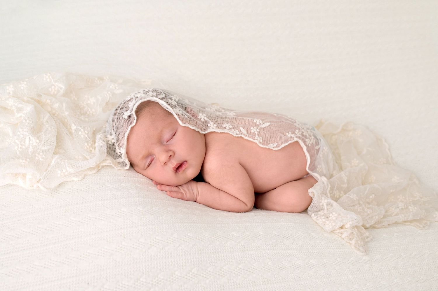 Sleeping newborn baby wearing a delicate lace bonnet on a soft white blanket.