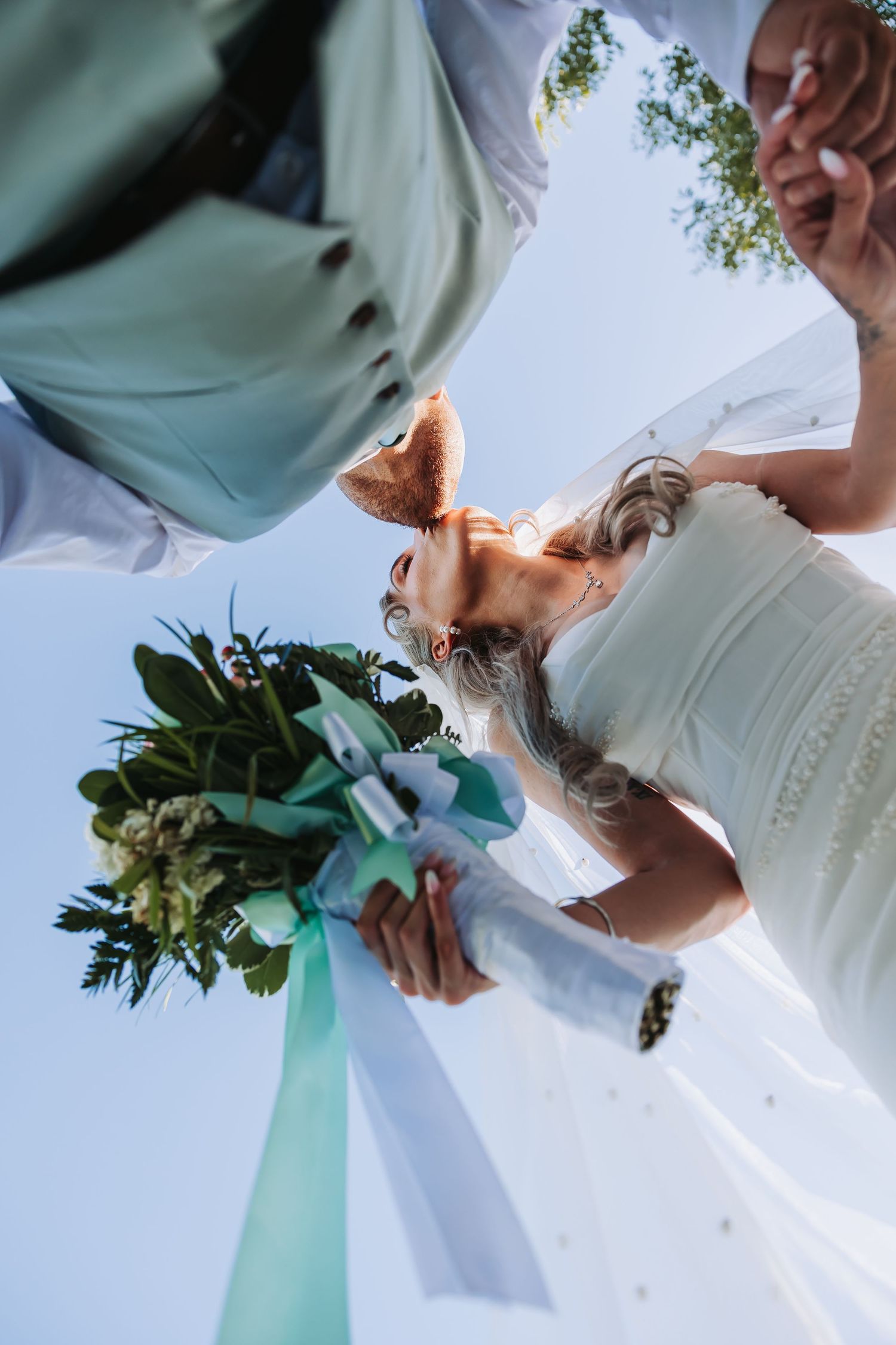 Bride and groom share a romantic kiss outdoors as wedding veil billows in wind, surrounded by white flowers and trees.