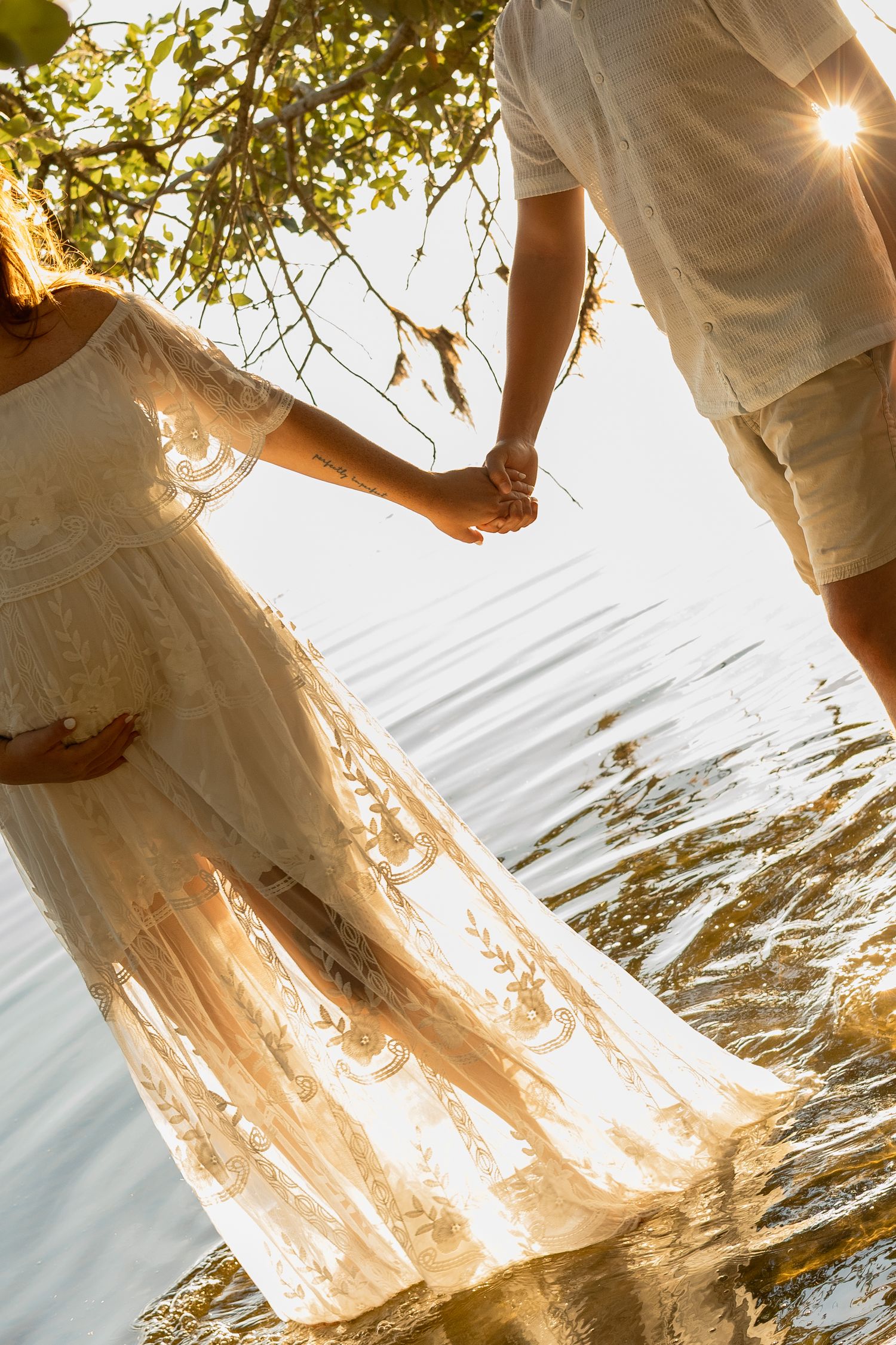 Couple holding hands in white outfits standing in water at golden sunset near tree branches.