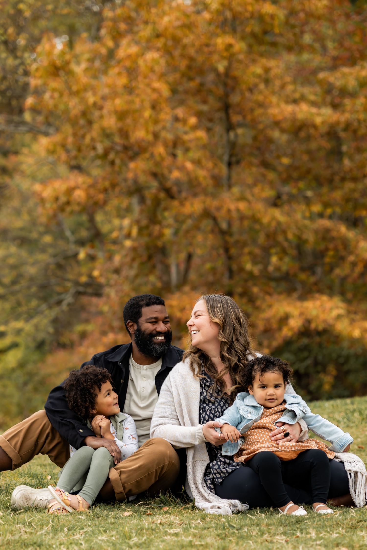 A happy interracial family sits together on autumn grass, smiling, with two young children and colorful fall trees behind them.