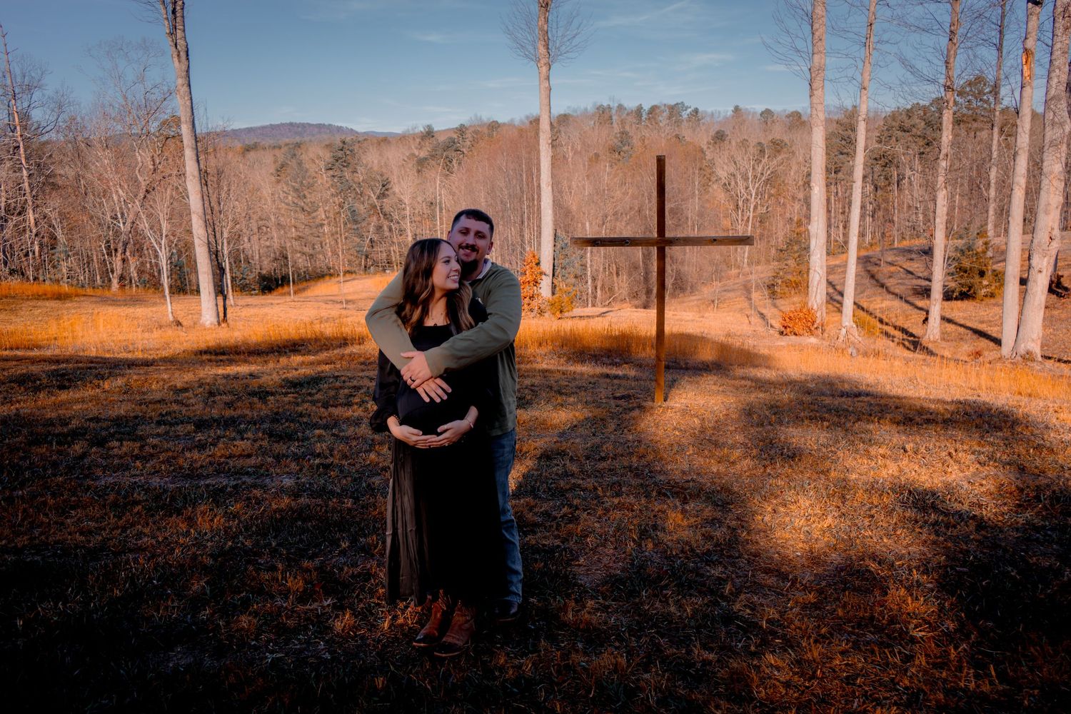 Couple sharing a tender maternity embrace near a wooden cross in a winter forest landscape.