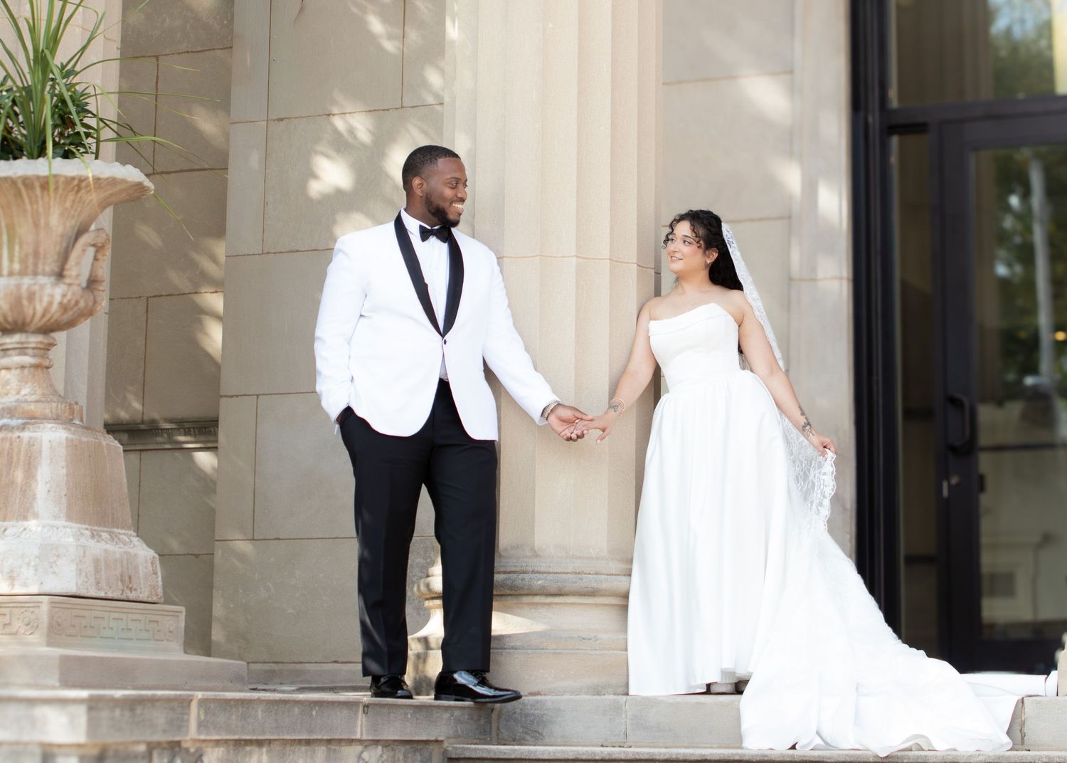 A bride holding white calla lilies and groom in a white tuxedo share a loving glance outdoors.
