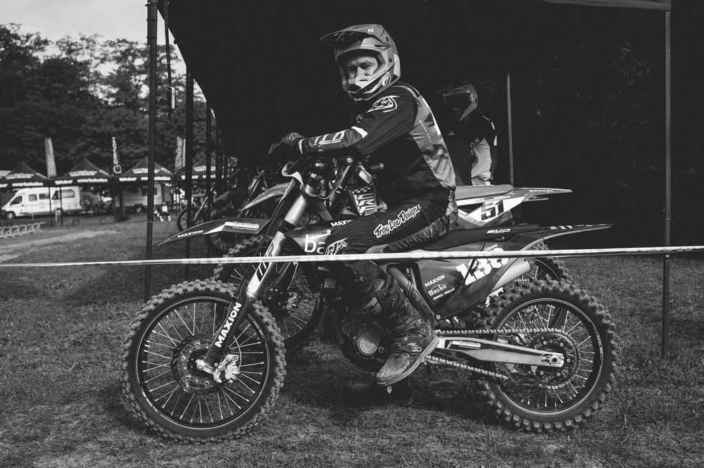Black and white photo of a motocross rider in full gear sitting on a dirt bike at an outdoor racing event.