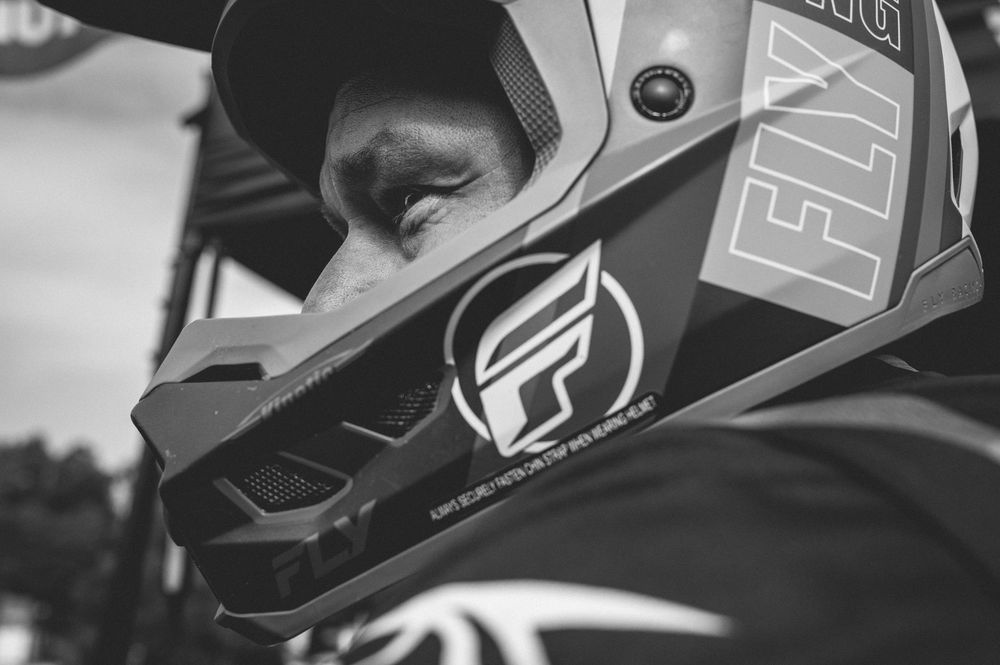 Black and white close-up of a motocross rider wearing a Fly Racing helmet at an outdoor event.