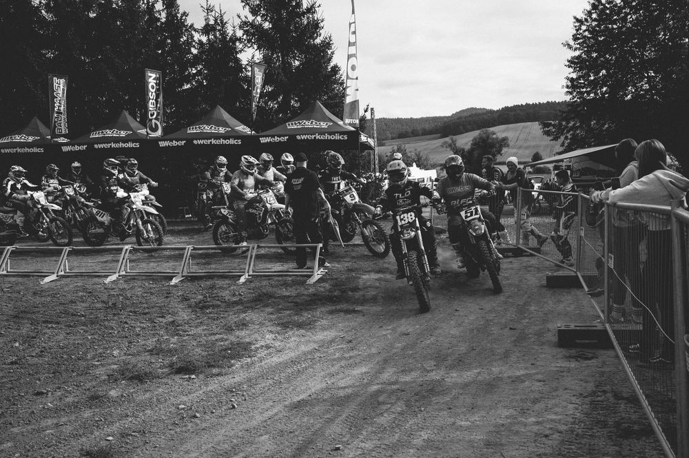 Black and white photo of motocross riders at the starting line of a race, with spectators and tents in the background.