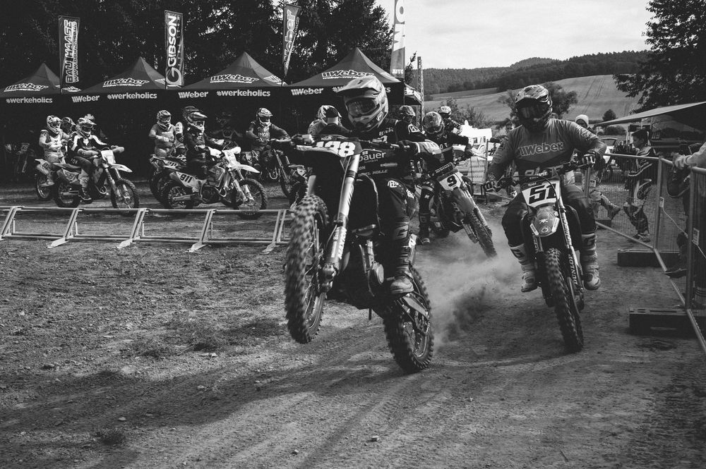 Black and white photo of motocross riders racing on dirt track with crowd watching in background.
