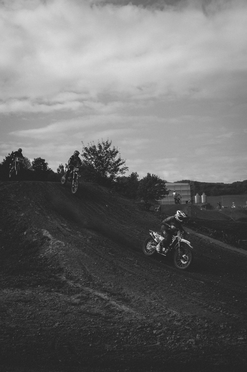 Black and white photo of motocross riders racing on a dirt track with trees and cloudy sky in the background.