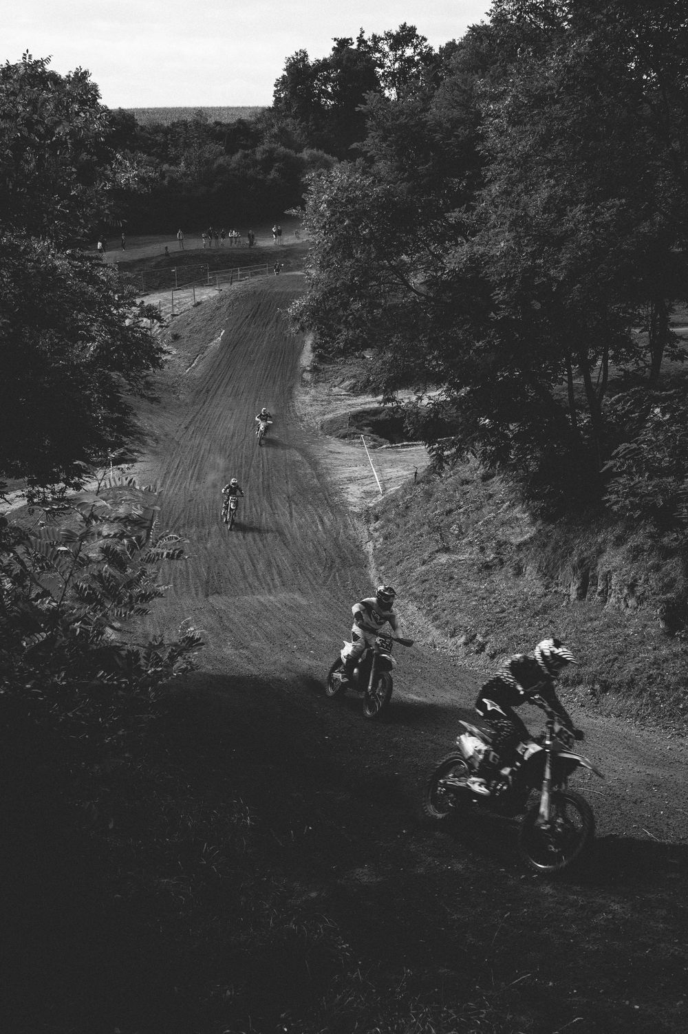 Black and white photo of motocross riders racing on a dirt track surrounded by trees.