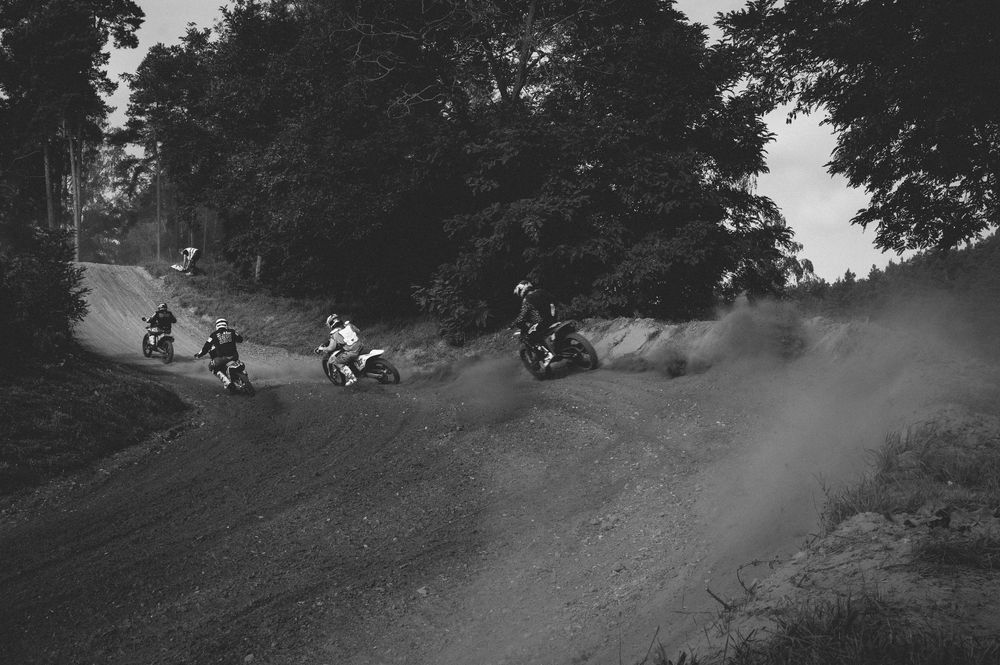 Black and white photo of motocross riders racing on a dusty dirt track surrounded by trees.