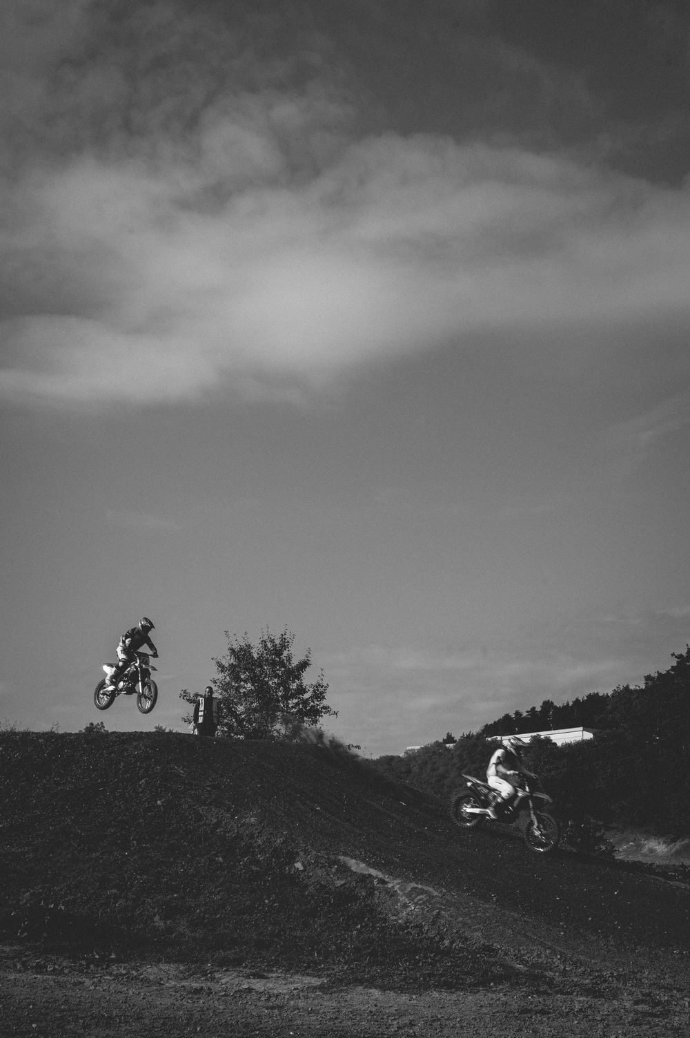 Two motocross riders perform jumps on a dirt track hill against a dramatic cloudy sky in black and white.